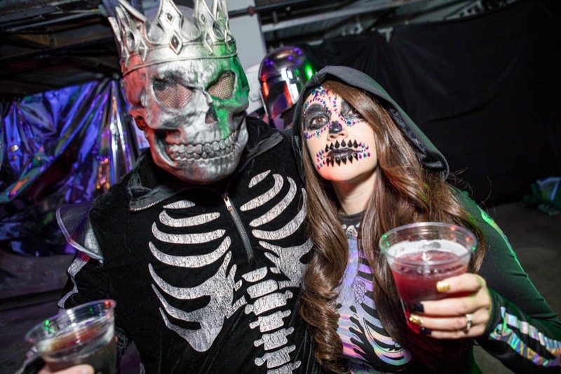 Crowned skull king and jeweled sugar-skull partner raise their drinks at the adults-only Coloween Halloween event