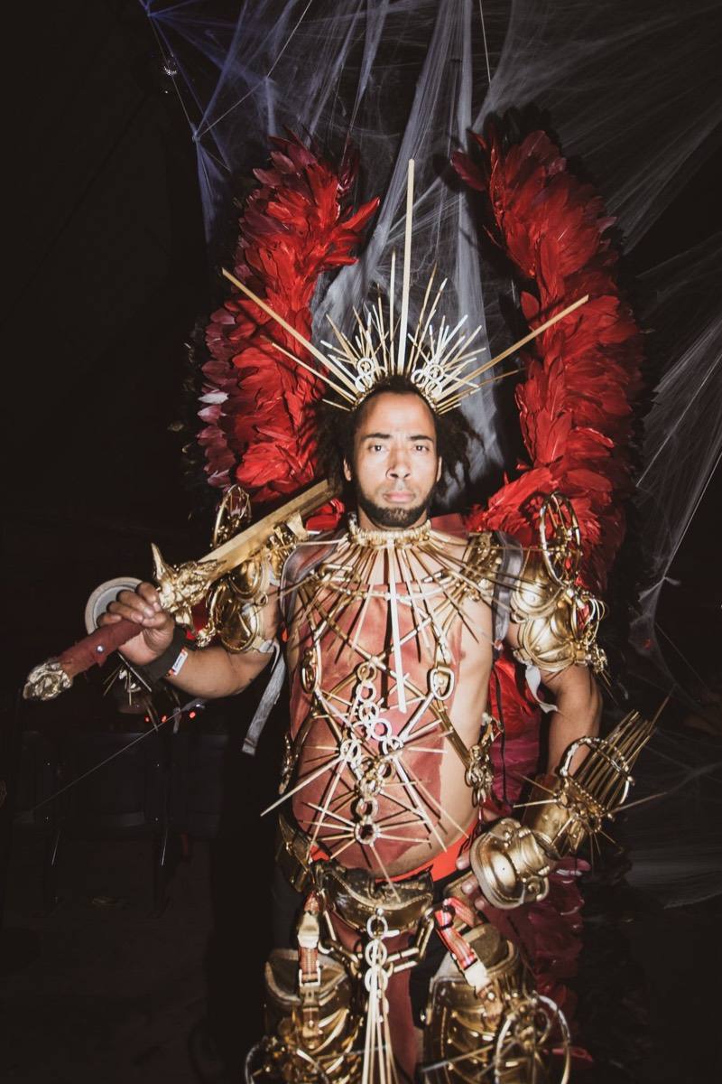 Winged warrior in gold armor and red feathers stares down the lens on the dance floor at Coloween 2022