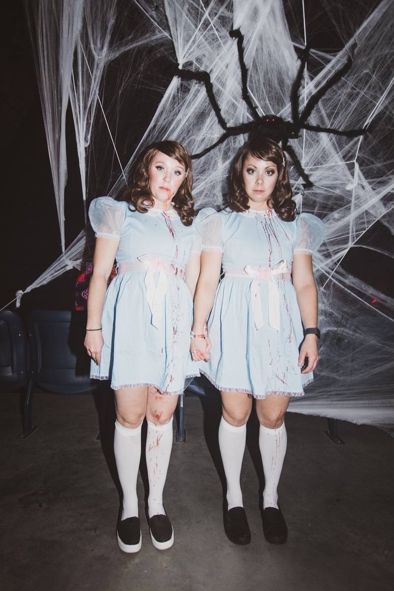 Twin blue dresses holding hands beneath a giant spider web at the adults-only Coloween Halloween event