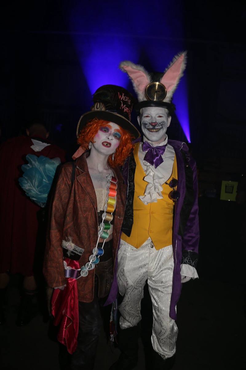 Mad Hatter and pocket-watch rabbit couple under blue beams at the adults-only Coloween Halloween event